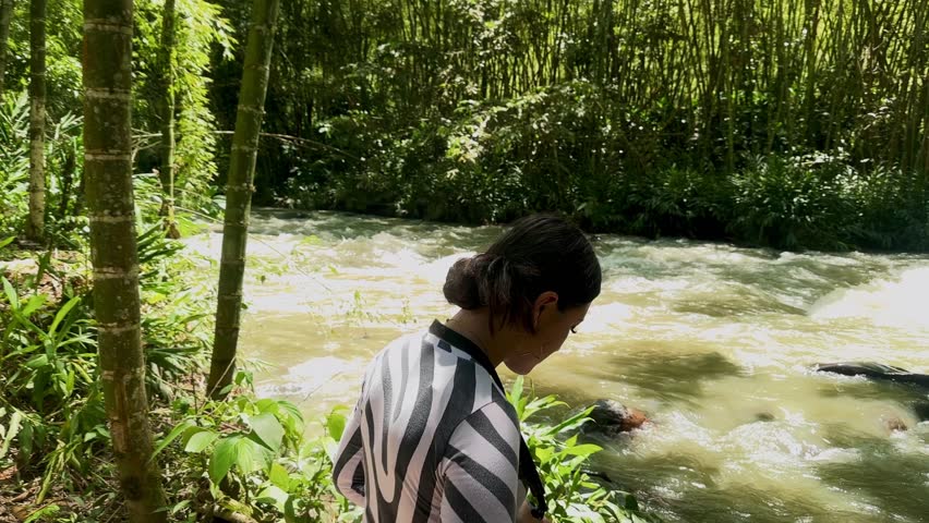 Colombian woman with black hair standing by the river during sunny vacation afternoon in El Santuario, Risaralda, holding camera and surrounded by tropical light and shadows
