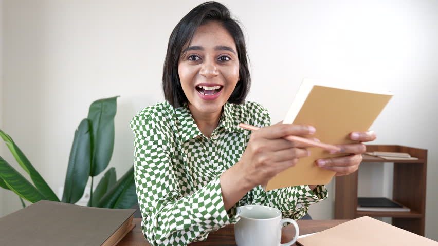 A happy woman with short dark hair, wearing a checkered shirt, holds a notebook and pencil, smiling widely with excitement