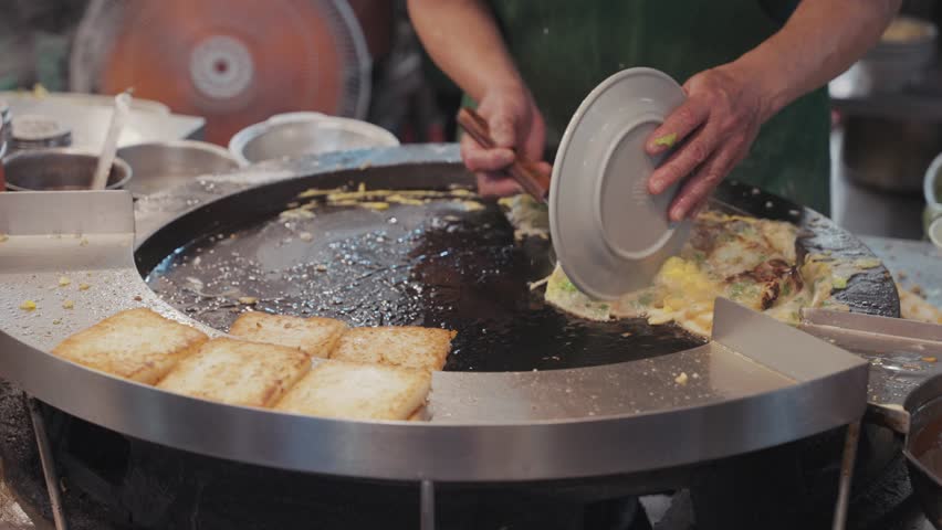 Street vendor frying and flipping sizzling Taiwanese oyster omelette on a hot round griddle, with eggs, oysters, and batter bubbling together into a savory street food delicacy.