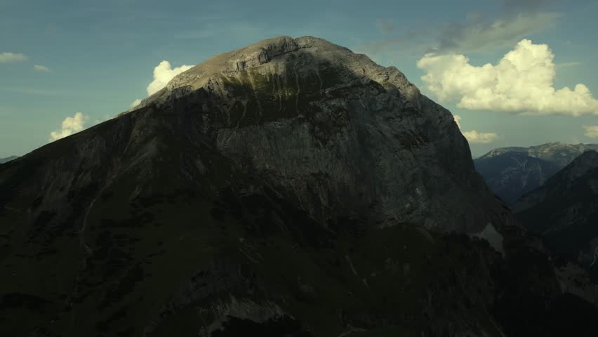 Aerial view of towering, rugged mountain peaks, their rocky faces contrasting with the green slopes under a sky dotted with fluffy clouds, Tirol, Austria.
