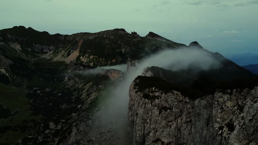 Aerial view of imposing mountain peaks, with dramatic cliffs and lush green vegetation, cloaked in ethereal mist, Klobenjoch, Tirol, Austria.