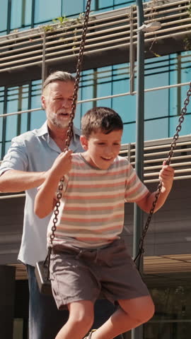 Vertical video, A grandfather pushes his grandson on a swing set at a playground. They are both laughing and having fun. The grandfather is wearing a light blue shirt and dark pants