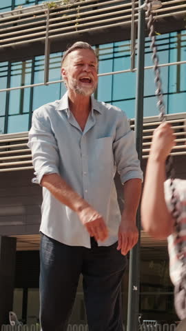 Vertical video, A grandfather pushes his grandson on a swing set at a playground. They are both laughing and having fun. The grandfather is wearing a light blue shirt and dark pants