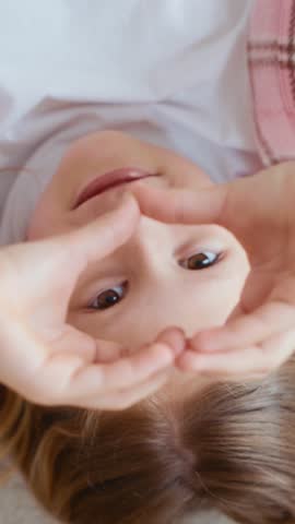 Girl lies on her back and forms a heart shape with her hands, looking through it into the camera. Joyful and expressive moment. Shot in vertical format, ideal for mobile or kids' content. 4k footage