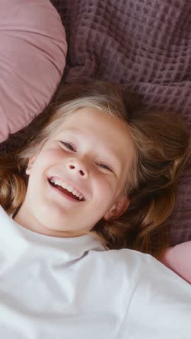 Girl falls back onto a bed full of soft pillows and smiles warmly into the camera. Cozy, carefree moment. Shot in vertical format, perfect for lifestyle or kids content. High quality 4k footage