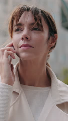 Vertical video, A young woman smiles while talking on her phone outdoors. She walks along a brick pathway past some trees and shrubbery.