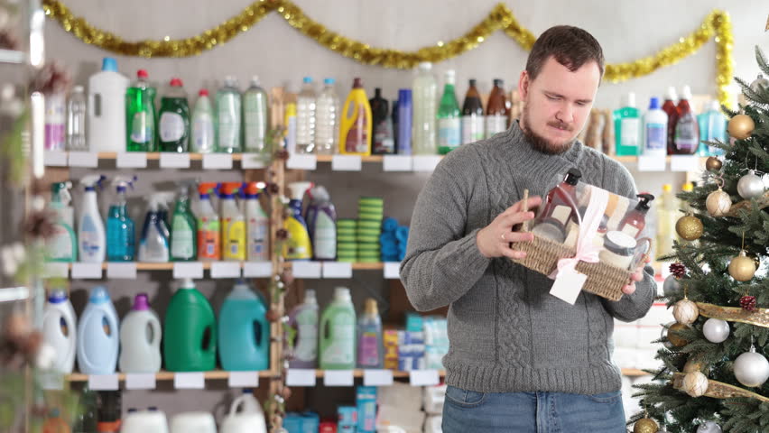 In Christmas decorated shop, man examines product and doubts, shower cosmetics present kit, study items properties in household department. Buyer examines and selects product.
