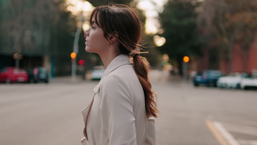 A young woman walks down a city street at sunset. She is wearing a stylish trench coat and has her hair tied back in a ponytail.