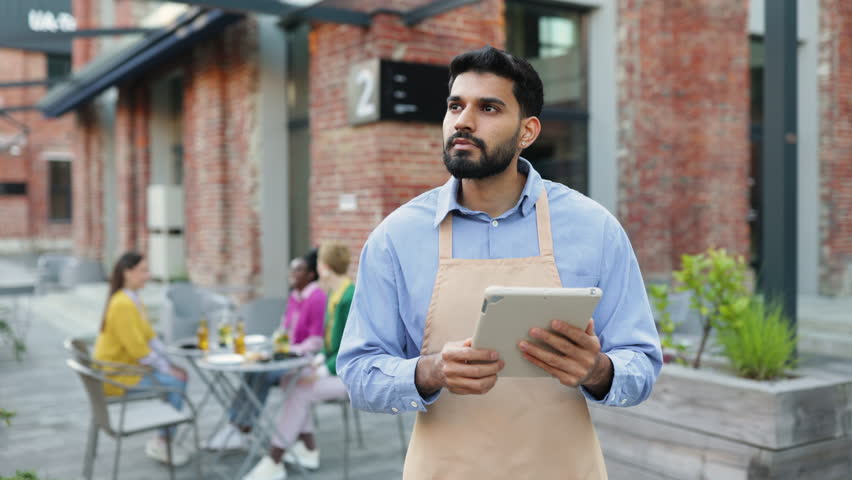 A Hindu male waiter holds a tablet, looking away from the camera at an outdoor restaurant. People sit at a table in the background.