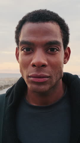 Vertical video, Young man stands at the beach during sunset. He looks directly into the camera with a serious and calm expression.