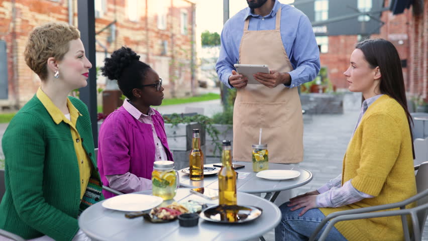 A diverse group of people enjoying a meal outdoors. A waiter takes an order on a tablet.