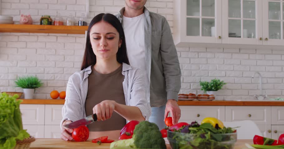 Happy young man having fun putting tomatoes on eyes of girlfriend. Couple preparing healthy salad with vegetables in kitchen at home. Food and nutrition concept. 4k video. Slow motion video.