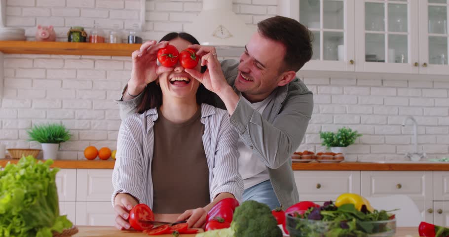 Happy young man having fun putting tomatoes on eyes of girlfriend. Couple preparing healthy salad with vegetables in kitchen at home. Food and nutrition concept. 4k video. Slow motion video.