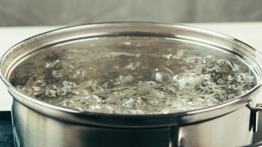 The chef in the restaurant is cooking while using the dipper in a large pot. The water is boiling and the mass of steam reflected in the morning light.