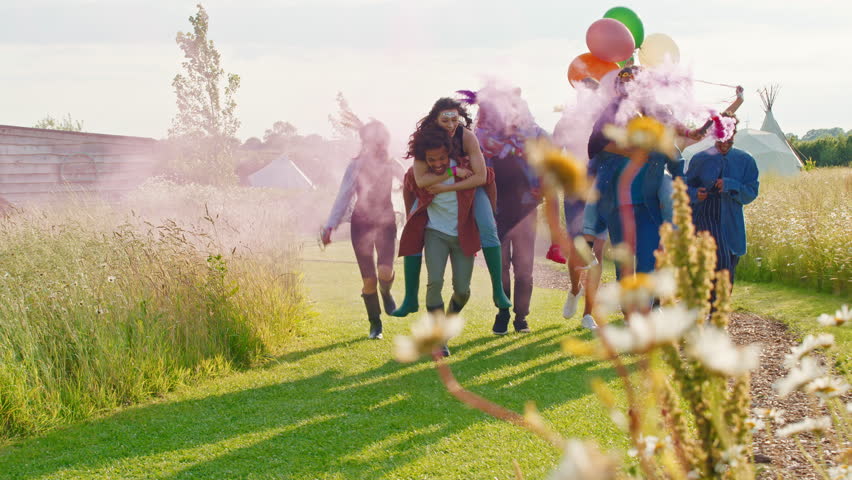 Group of young friends walking through music festival site giving one another piggyback rides carrying smoke flare and balloons - shot in slow motion