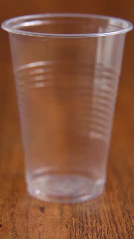 Clear water stream begins pouring into empty transparent plastic cup on wooden surface. Water flows steadily into plastic cup. Male hand reaches to grasp filled plastic water cup from wooden surface.