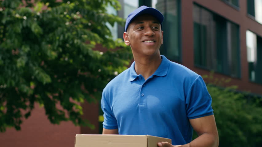 Smiling positive African American ethnic courier delivery man male guy post worker holding parcel carrying postal order shopping service deliver walking in city wearing blue uniform walk outdoors job