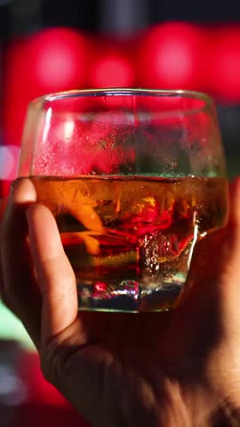 A hand slowly rotates a short glass of whiskey with ice in front of vivid red and green bar lights, creating a dramatic, cinematic atmosphere