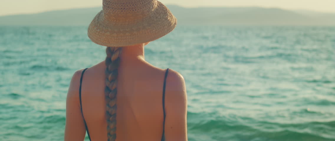 Woman with braid and straw hat looking at sea horizon in summer sunny day