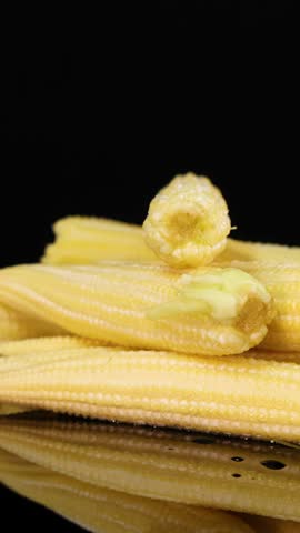 A pile of yellow baby corn rotates smoothly on a reflective black surface, illuminated by soft studio lighting, highlighting texture and freshness in close-up detail