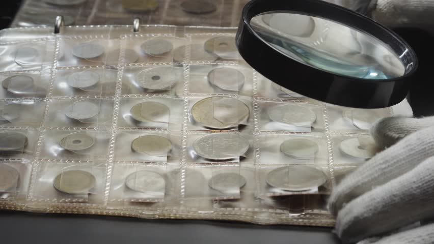 Coin collector uses magnifying glass and gloves to examine collection