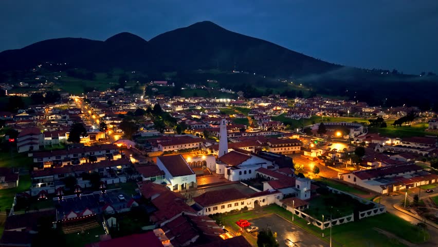 Guatavita, Colombia at dusk, with warm village lights glowing beneath a looming dark mountain backdrop.