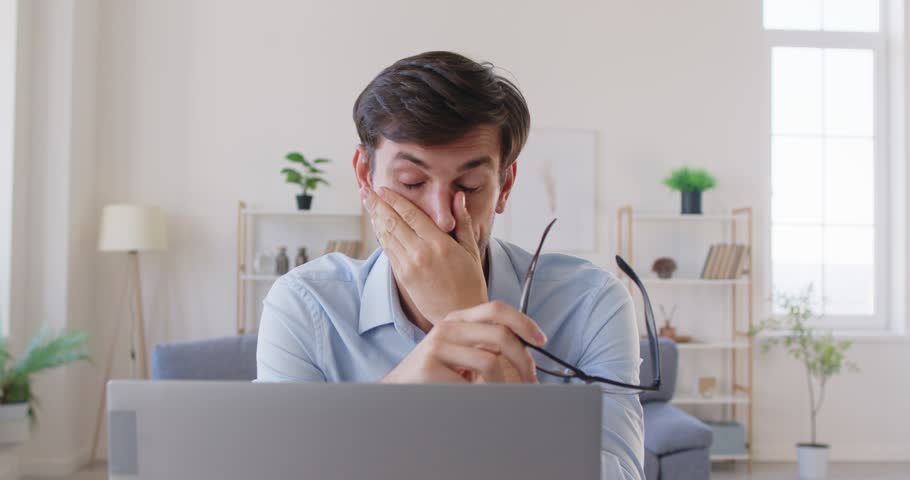 Tired young overworked business man suffering from headache after long hours computer work and taking off glasses sitting at desk on workplace in office in front of laptop. 4k video.