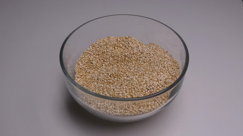 A clear glass bowl filled with raw uncooked white quinoa. A hand comes into frame and dips its fingers into the quinoa and starts touching and inspecting the grains.
