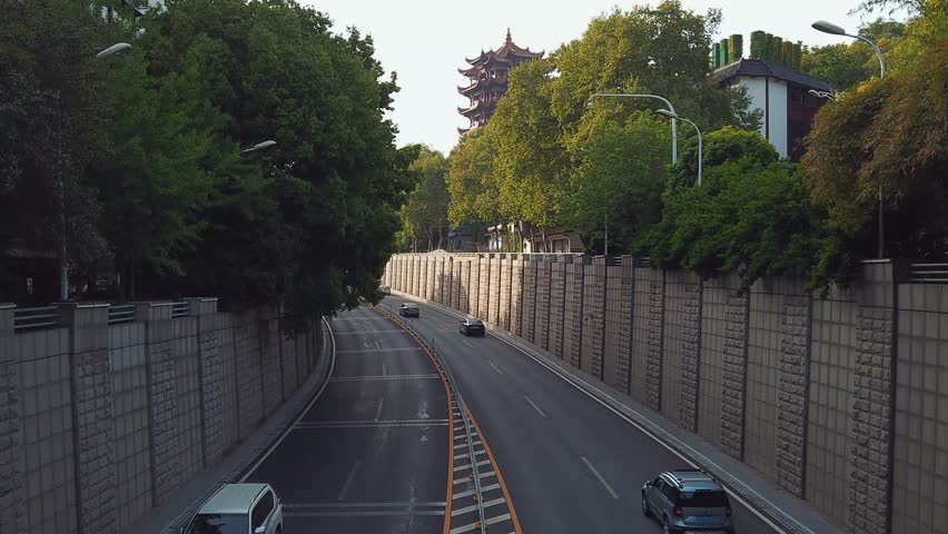 The bustling road near Huanghe Tower in Wuhan is captured in a captivating timelapse, displaying the dynamic flow of traffic against a backdrop of green trees and traditional architecture.