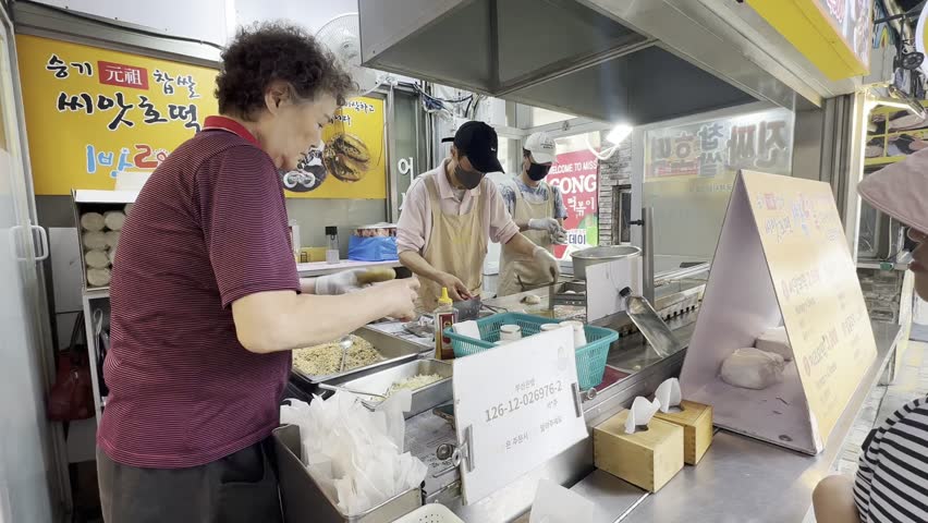 Busan, South Korea – June 15, 2025: Street food vendors prepare hotteok, a popular Korean stuffed pancake, using scissors and tongs at a bustling market food stall.

