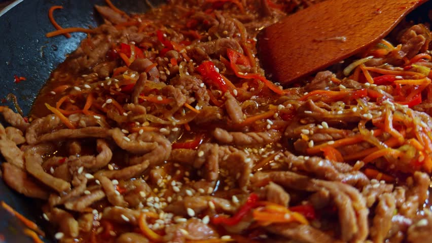 stir-fried pork with julienned carrots and bell peppers in a savory sauce, topped with sesame seeds. Hot dish being cooked in wok with wooden spatula, Asian-style home cooking. Thai food exotic flavor