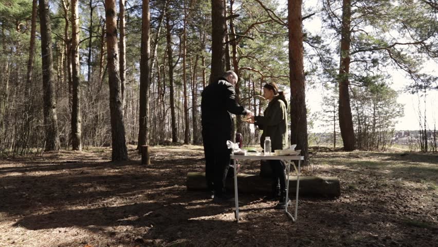 Two people camping in forest glade, preparing drinks and snacks.