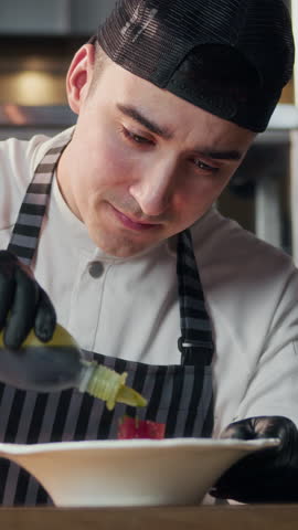 Vertical shot of young chef in uniform flavoring fish dish on plate while working in modern restaurant kitchen