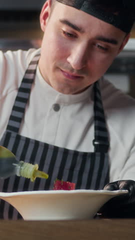 Vertical shot of young chef in uniform flavoring fish dish on plate while working in modern restaurant kitchen