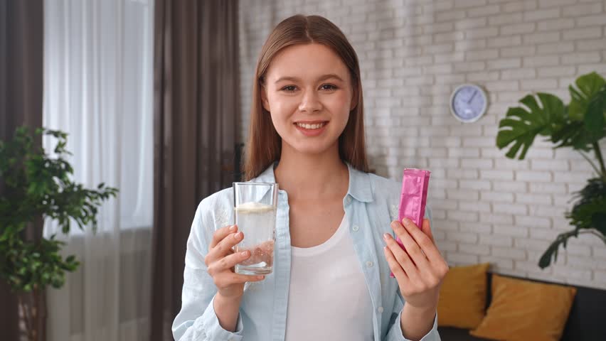 A young woman pours powder collagen into a glass of water, smiling and preparing a refreshing drink in a modern kitchen. Healthy living concept, taking vitamins for skin and hair beauty.