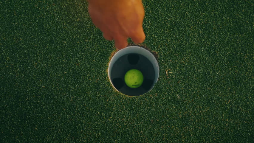 Top-down slow motion shot of a yellow golf ball rolling into the cup, bouncing inside the hole, while a male golfer’s arm reaches in to retrieve it. A close, dynamic moment capturing golf precision.