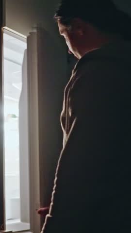 A person is peering into an open refrigerator at night, considering latenight snacks