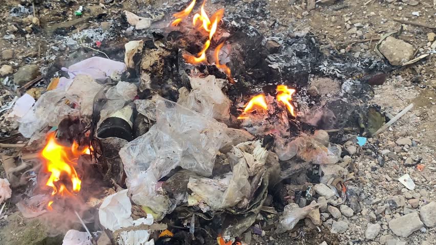 A pile of burning plastic trash on a dirt ground releases smoke containing harmful chemicals and carcinogens into the air, posing significant health risks, including cancer
