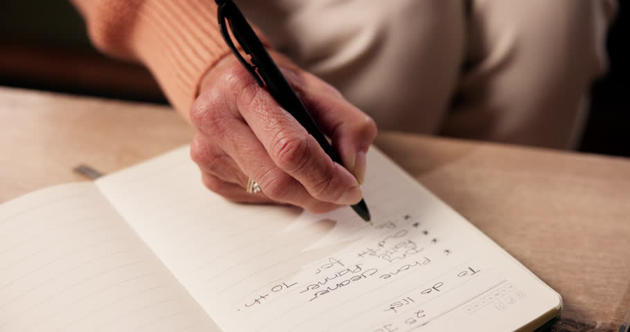 Hand, to do list and writing with person in living room of home for planning or reminder of tasks. Diary, journal or notebook with senior woman in apartment for agenda, checklist or event schedule