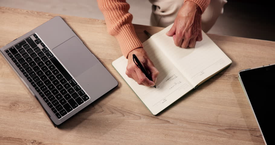 Hand, laptop and notebook with person in living room of home for planning or reminder of tasks. Computer, journal and writing with senior woman in apartment for agenda, checklist or event schedule