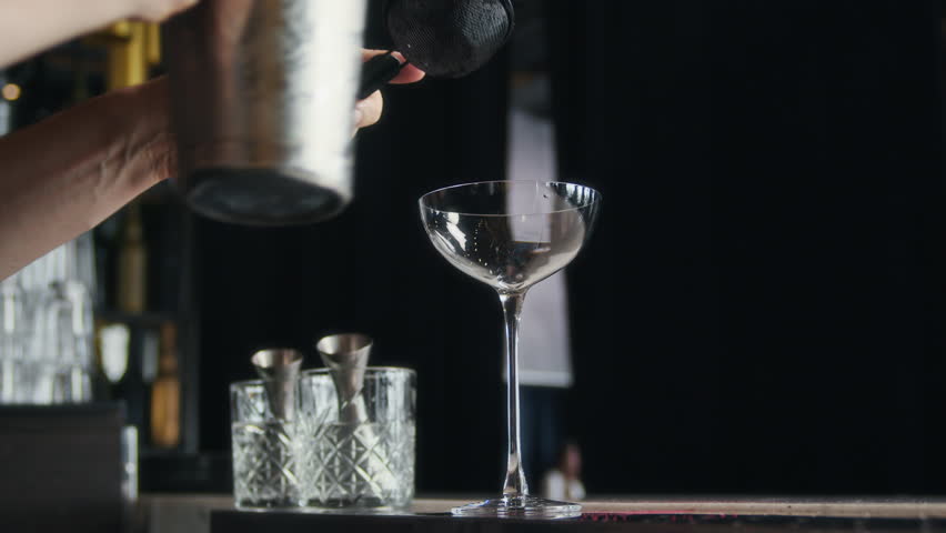 Cropped shot of hands of unrecognizable barman pouring cocktail in glass through strainer at bar counter