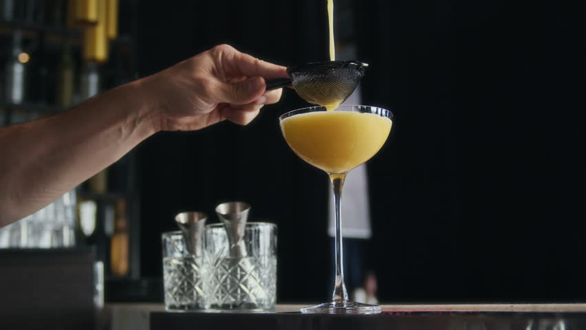Cropped shot of hands of unrecognizable barman pouring cocktail in glass through strainer at bar counter