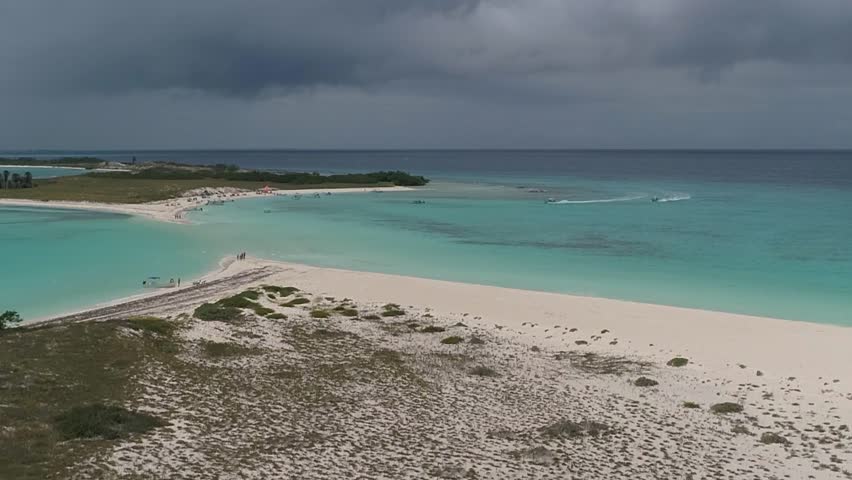 Aerial rise over Cayo de Agua, Caribbean island in Los Roques, Venezuela, with boats crossing shallow turquoise water toward white sands, where people enjoy a sunny beach paradise.