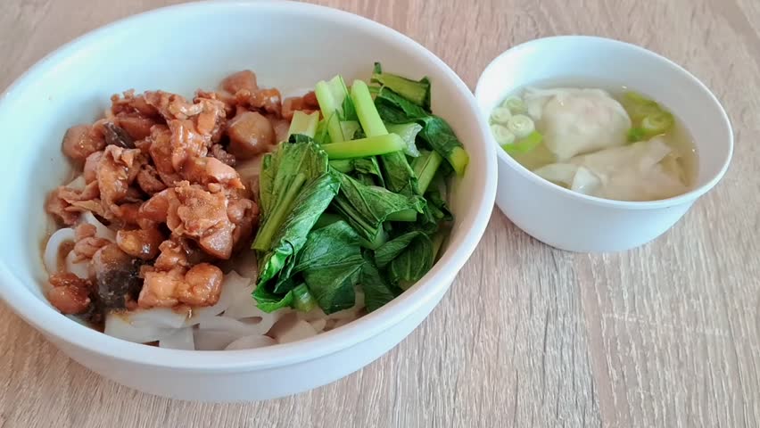 A closeup view of a bowl of noodle (kwetiau) with bok choy vegetables and chicken meat; and a mall bowl of dumplings soup.