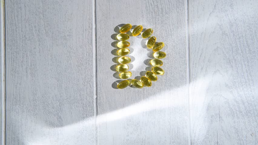 Yellow transparent capsules of vitamin d forming the letter d on a white wooden surface illuminated by sunlight, representing the importance of this vitamin for health and well-being