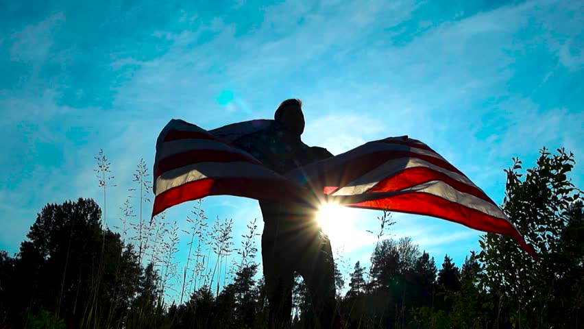 Patriotic man that looks like a silhouette waving the United States of America flag in slow motion in nature, filmed from a low angle and sun is shining behind the flag and the man that are moving.