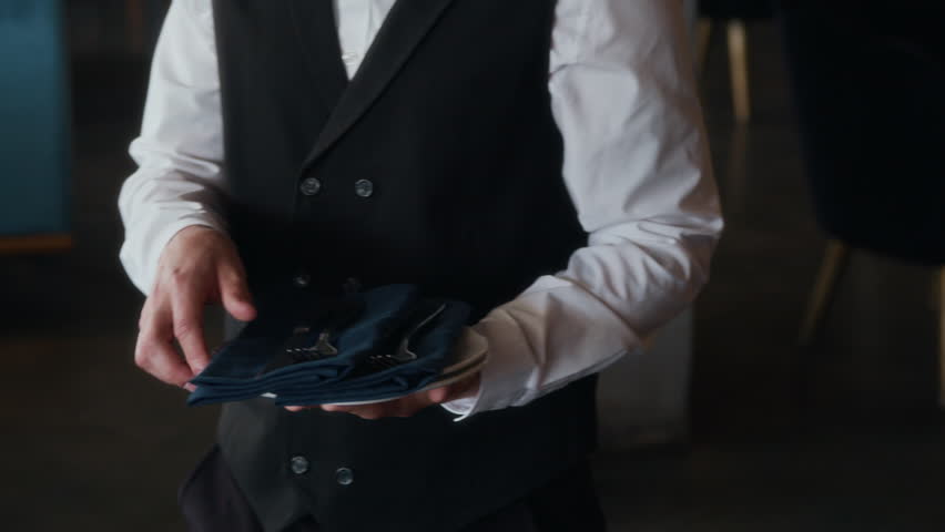 Cropped shot of hands of unrecognizable waiter in black and white uniform serving plates and cutlery on table in restaurant