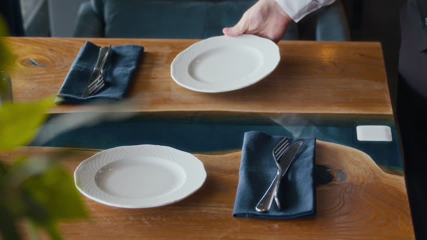 Cropped shot of hands of unrecognizable waiter in black and white uniform serving plates and cutlery on table in restaurant