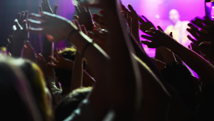 Concert crowd enthusiastically waving hands, recording live music performance under vibrant purple stage lights, capturing electrifying moment of shared musical experience and excitement