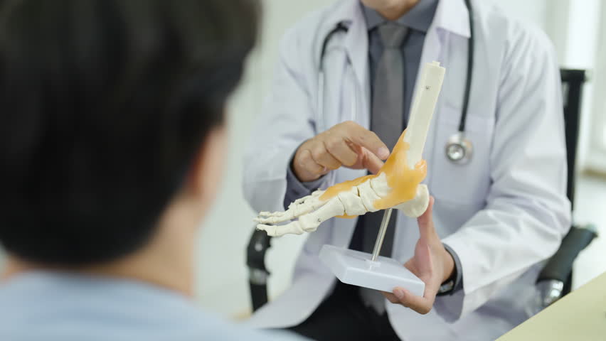 Doctor explaining foot joint anatomy to patient using realistic bone model during orthopedic consultation, healthcare professional demonstrating bone structure and discussing foot or ankle condition.